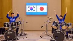 李氏と高市氏がそれぞれ、ドラムセットに座っている。共に青いジャケットを着て、両手に持ったドラムスティックを掲げ、笑顔を見せている。ドラムセットの間に設置されたモニターに、韓国と日本の国旗が並んで表示されている