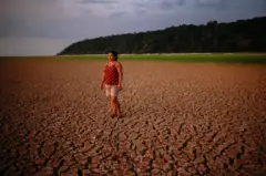 Uma mulher de pé no leito seco e rachado de um rio, com um fio de água ao longe
