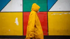 A man in a yellow rain coat walks past a brightly coloured mural.