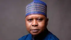 Kabiru Taminu Turaki wọ aṣọ olómi aro, dé fìlà, tó sì ń wo ẹ̀rọ ayàwòrán 