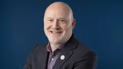 Homem com barba por fazer branca no queixo sorri para a câmera. Ele está vestindo uma camisa e um paletó de tweed. O fundo é uma tela fotográfica azul.