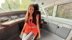 Молода дівчина із довгим каштановим волоссям, вдягнена у яскравий помаранчевий сигнальний жилет, сидить у кріслі машиніста поїзда. Навколо видно панелі керування, екрани й великі вікна, через які проглядаються дерева зовні.