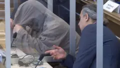 شخص يتكلم مع إياد غريب في المحكمة وإياد غريب يرتدي قميصا رمادي اللون ويغطي وجهه بقبعة القميص