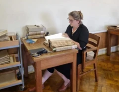 Pesquisadora Alícia Duhá Lose sentada em uma sala de arquivo, analisando um manuscrito antigo sobre a mesa. Ao redor, há pilhas de livros e documentos históricos, em ambiente de pesquisa na Universidade de Coimbra