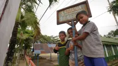 Tiga anak berpose di plang Jalan Kebudayaan. Plang jalan itu ditulis dalam dua aksara, yakni aksara latin dan Hangeul.