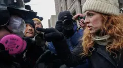 A woman with red hair and wearing a white cable-knit beanie hat points a gloved finger at a man dressed in googles, a hardhat and a pink respirator mask. She looks angry but not afraid. the action is happening in a crowded group of people, a cameraman is taking photographs behind them.