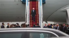 Lula descendo de avião em tapete vermelho, com várias pessoas à sua espera na pista