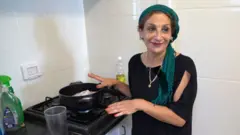 Uma mulher está ao lado de uma panela preta aberta no fogão. Ela usa um lenço verde na cabeça, blusa preta e um colar dourado com uma estrela. Há uma garrafa de óleo atrás dela e as paredes são revestidas com azulejos brancos.