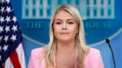 La secretaria de Prensa de la Casa Blanca, Karoline Leavitt, en la sala de prensa. Viste una chaqueta rosa y tiene detrás la bandera de EE.UU. y el cartel de The White House