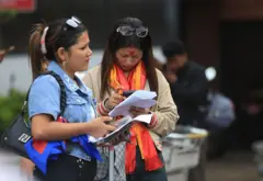 काठमाण्डू विमानस्थलमा विदेश जाने तरखरमा रहेका दुई युवतीहरू, एक जनाले रातो टीका र खादा लगाएर मोबाइल फोन चलाइरहेकी छिन् भने अर्कीले नेपालको झण्डा भिरेर कागजपत्र लिएकी छन्