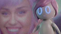 Imagem de personagem interpretada por Miley Cyrus aparece em tela e, na frente, a robô Ashley
