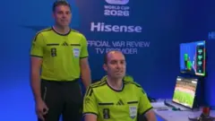 O árbitro Shaun Evans, à esquerda, faz com as mãos um OK de cabeça para baixo no centro de arbitragem da Fifa.