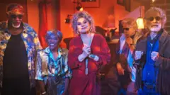 Um grupo de cinco idosos está reunido em um ambiente com iluminação quente que lembra um bar ou lounge: à esquerda, Tony Tornado, de pele escura com óculos escuros, faixa na cabeça e camisa estampada; ao lado, Teca Pereira, de pele escura, sorridente, com cabelo curto grisalho; no centro, Vera Fischer, de pele clara, com cabelo volumoso e vestido vermelho; ao fundo, Reginaldo Faria, de pele clara, com óculos escuros, sentado; e à direita Hamilton Vaz Pereira, de pele clara, com cabelo e barba grisalhos, também de óculos escuros, vestindo camisa jeans e jaqueta com franjas, com as mãos erguidas.