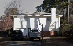Imagem mostra fachada de uma casa branca, com dois andares, dois carros, um preto e outro branco, em frente à casa.