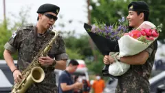 RM, Kim Namjoon is wearing his military uniform and sunglasses, he is playing the saxophone. V, Kim Tae-hyung, is looking at RM, also clad in military uniform, holding two bouquets of purple and pink flowers.