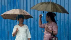 ရန်ကုန်မိုးကို အံတုပြီး ကားမှတ်တိုင်မှာ ကားစောင့်နေကြတဲ့ အမျိုးသမီးတွေကို ၂၀၂၅ ခုနှစ် ဇူလိုင်လတုန်းက မြင်တွေ့ရတာဖြစ်ပါတယ်။​ အမျိုးသမီးနှစ်ဦး ထီးကိုယ်စီ ဆောင်းထားကြပြီး သူတို့အနောက်မှာ အပြာရောင် သွပ်ကာထားတဲ့ ဆောက်လုပ်ရေးလုပ်ငန်းခွင်ရှိနေ 