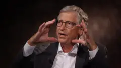 Bill Gates bir televizyon röportajında ellerini kaldırmış şekilde bir şeyler anlatıyor. Üzerinde kravatsız bir takım var