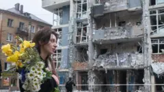 Kobieta z bukietem kwiatów przechodzi obok wysokiego budynku mieszkalnego w dzielnicy Szewczenkowskiej w Kijowie 25 maja 2025 roku. Jej włosy częściowo opadają na czoło. Za nią, na ulicy, stoi wielopiętrowy budynek poważnie uszkodzony wskutek ataku rosyjskich dronów.