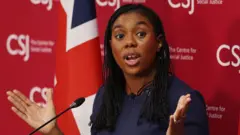 Kemi Badenoch dey wia blue cloth and tok for front of podium wit red branded background