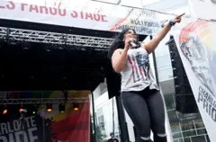 A cantora Vassy aponta para o público a partir de um palco. Ela usa uma camiseta branca com uma bandeira americana estampada. Atrás dela, há um palco com uma faixa arco-íris que diz “Orgulho gay Charlotte”.