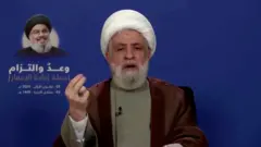 نعیم قاسم با عکس حسن نصرالله در پشت سرش در حال سخنرانی که از تلویزیون پخش می‌شود.