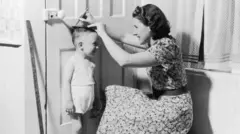 Мати позначає олівцем зріст свого сина на дверях, приблизно 1950 рік. Це чорно-біле фото, на якому видно посмішки матері та дитини. Жінка одягнена в квітчасту сукню, а дитина — у майку та штани.