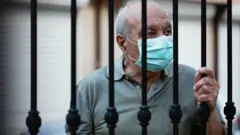 Imagem de banco de imagens mostra homem idoso com máscara olhando através de grade