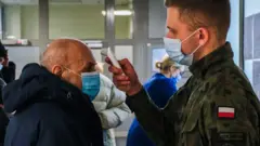 A military man wears a protective face mask and gloves as he screens the temperature of an elderly man before being given the Pfizer/BioNTech COVID-19 jab at the Krakow University Hospital on 25 January 2021 in Krakow, Poland