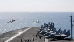 Yedi savaş uçağı, uçak gemisinin güvertesinde park halinde duruyor. İki uçak henüz kalkmış durumda ve deniz üzerinde yükseliyorlar.