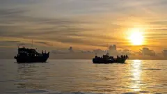 Two boats carrying lots of people float in a sea against a setting sun
