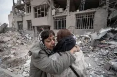 دو زن در آغوش هم در حالیکه پشت سرشان خانه‌ای ویران شده قرار دارد