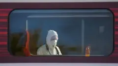 Cabeça e ombros de um perito policial são vistos através da janela de um vagão de trem. Ele usa capuz branco sobre a cabeça e máscara branca cobrindo nariz e boca.