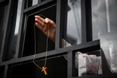 Pessoa em janela segurando colar com cruz de madeira