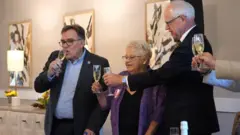 Anita LeBrun usa um blazer roxo e óculos enquanto ergue uma taça de champanhe para brindar ao governador de Minnesota, Tim Walz — que é mais alto e calvo, e segura sua própria taça próxima à dela.