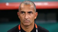 Ifoto ngufi igaragaza Sabri Lamouchi ahagaze mu gace kagenewe abatoza ku kibuga. Arareba muri 'camera' asa n'ureba hejuru. Yambaye umwenda w'umukara. Mu ijosi yiteye umushumi uriho ibara rya 'orange' n'umweru.