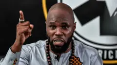 Kemi Seba, vêtu d'une chemise grise, est de face. Il porte un collier de perles en bois et son bras gauche est levé, l'index pointé vers le haut. À l'arrière-plan, on aperçoit partiellement le logo de l'ONG Pan-Africanist Emergency.