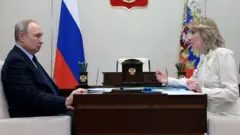 Vladimir Putin e Maria Lvova-Belova sentados em lados opostos de uma mesa, com uma bandeira russa ao fundo