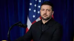 青いカーテンと星条旗を背にしたゼレンスキー氏。黒い上着とシャツを着て、厳しい表情をしている