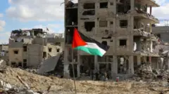 يرفرف علم فلسطيني قرب أنقاض المنازل والمباني المدمرة خلال الحرب، في أعقاب وقف إطلاق النار بين إسرائيل وحماس، في رفح في جنوب قطاع غزة، 20 يناير/كانون الثاني 2025. الصورة التقطت نهاراً.