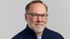 弗雷德·拉姆斯德爾 (Fred Ramsdell) 身穿海軍藍套頭衫，戴著黑框眼鏡，在自然背景前看著鏡頭。