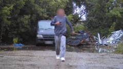 Photo de surveillance montrant un adolescent vêtu d'un survêtement gris, le visage flouté, tenant un téléphone portable. Il s'éloigne d'une camionnette grise stationnée sur une surface dure dans une zone boisée.