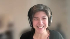 Uma mulher sorri enquanto usa fones de ouvido grandes, em um ambiente interno com fundo desfocado, em uma chamada de vídeo.