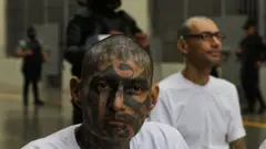 Un pandillero escucha una audiencia virtual durante un juicio masivo contra miembros de la pandilla Mara Salvatrucha (MS-13), celebrado en el Centro de Confinamiento por Terrorismo (Cecot), en el municipio de Tecoluca, San Vicente, El Salvador, el 23 de abril de 2026. Un detenido tiene una franela blanca y toda la cara tatuada.
