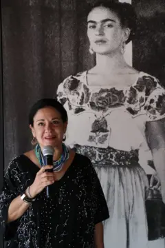 Cristina Kahlo Alcalá usa um vestido preto com bolinhas brancas, brincos e um colar azul e branco de várias voltas. Tem um microfone na mão e, atrás dela, há uma foto de Frida Kahlo olhando para a câmera. Frida está vestida com traje tradicional de tehuana