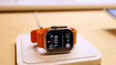 Apple Watch com mostrador preto e pulseira laranja sobre uma mesa, marcando 1h03