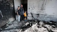 Un padre abraza a sus dos hijos mientras observa los restos de una casa incendiada. Hay escombros, tubos retorcidos y vigas caídas