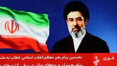 Ifoto yakuwe muri videwo yo mu makuru yo kuri televiziyo ya Irani, igaragaza ifoto ya Mojtaba Khamenei ari iruhande rw'ibendera rya Irani. Igice cyo hasi cy'ifoto kirimo amagambo yo mu rurimi rw'Igi-Farsi yanditse mu mweru ku mutuku. 