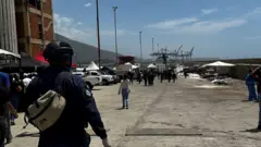 Un policía en primer plano de espaldas y al fondo se ven cuerpos tirados cubiertos con bolsas de plástico.