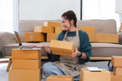 Una mujer sonriente, sentada en el piso, rodeada de cajas, consulta su computador portátil