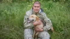 Andrey Spivak, sentado entre hierba alta, sostiene a un perro de color beige y negro. Viste un conjunto de exterior con estampado de camuflaje, lleva el pelo rapado al ras y mira directamente a la cámara.